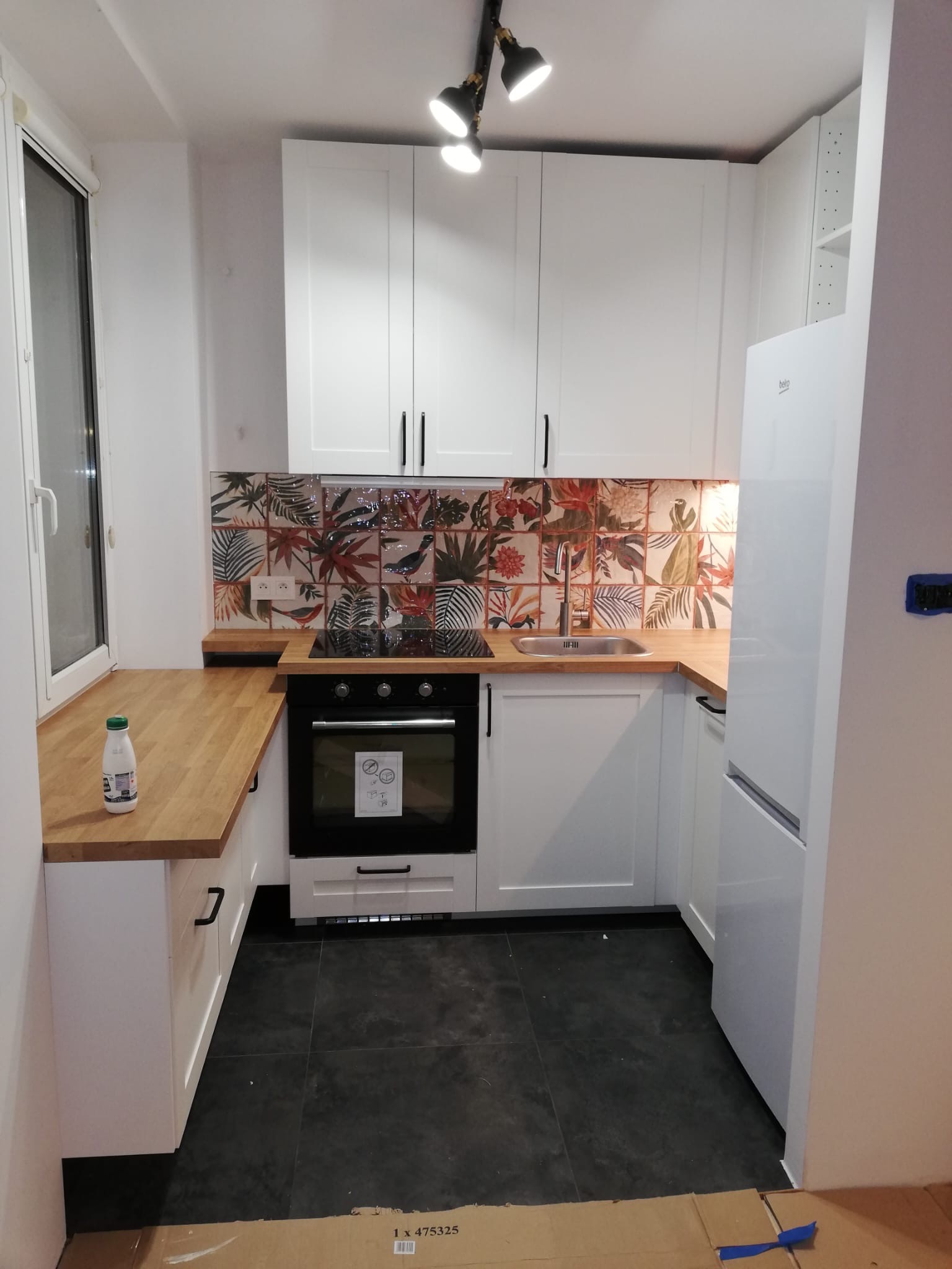 Nowoczesna, jasna kuchnia z białymi szafkami, drewnianym blatem i ozdobnymi płytkami nad blatem, czarna podłoga, widoczna butelka na blacie i karton na podłodze.