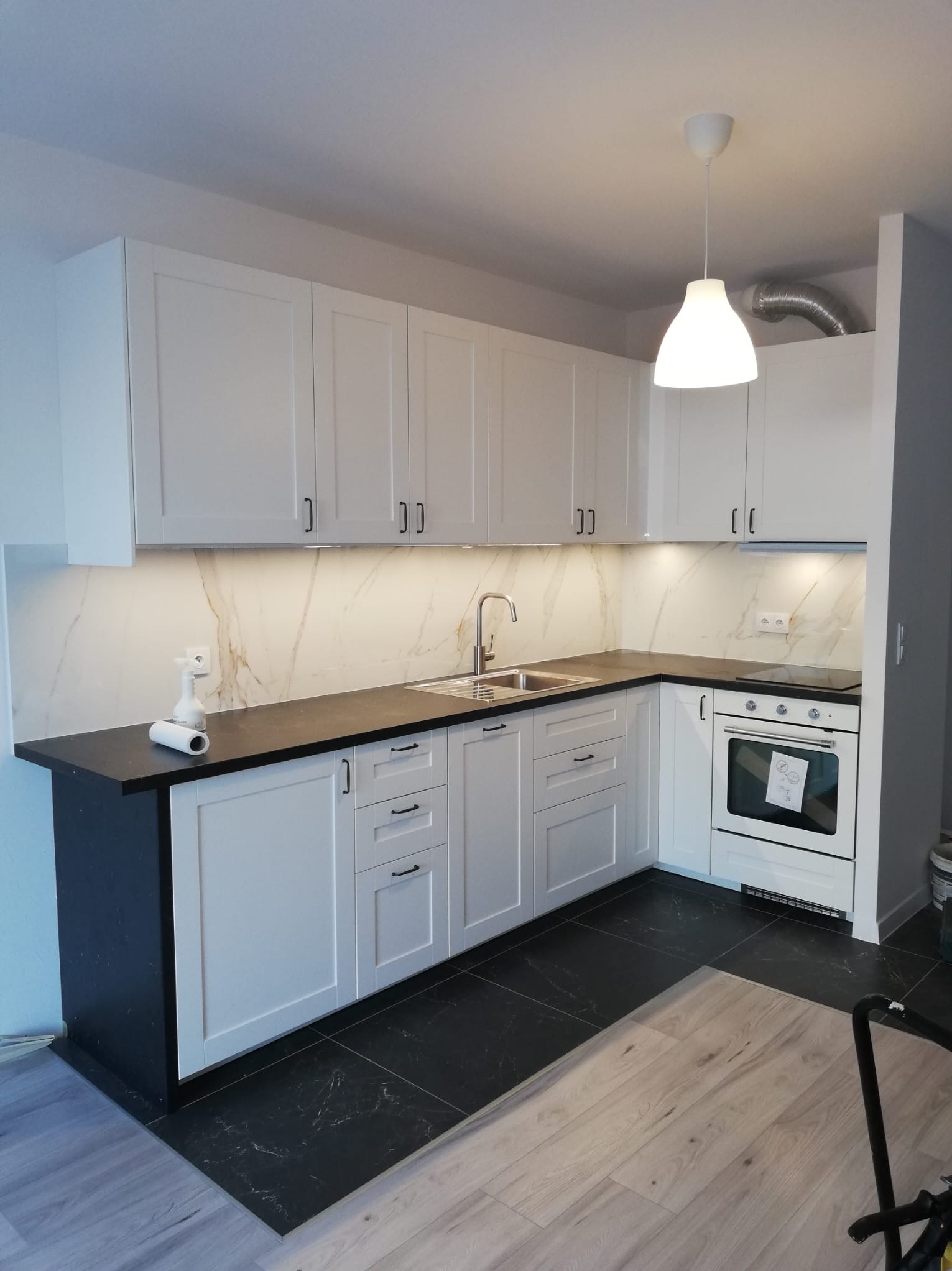 Nowoczesna, jasna kuchnia z białymi frontami szafek, czarnym blatem i marmurowym backsplashiem podświetlonym ledami, widok na zlewozmywak i piekarnik.