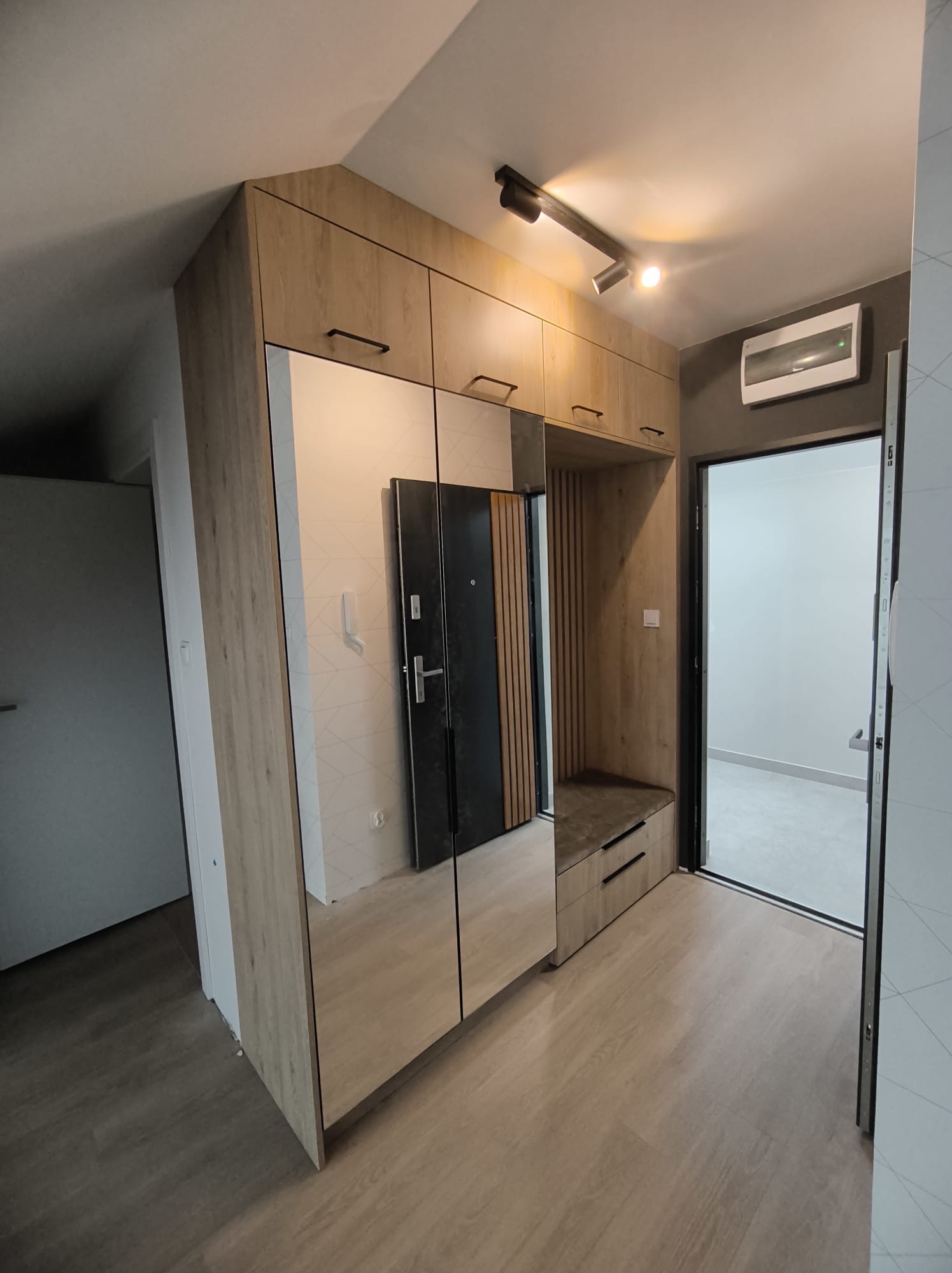 Hall zabudowany szafą z lustrami i siedziskiem w odcieniach drewna, oświetlenie punktowe na suficie, widok na drzwi wejściowe i wewnętrzne.