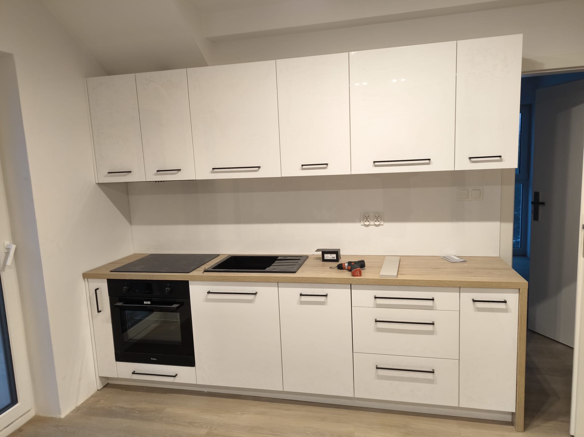 Biała zabudowa kuchenna z drewnianym blatem, czarnym piekarnikiem i płytą grzewczą, w trakcie montażu widoczna wiertarka i listwa przypodłogowa na blacie.
