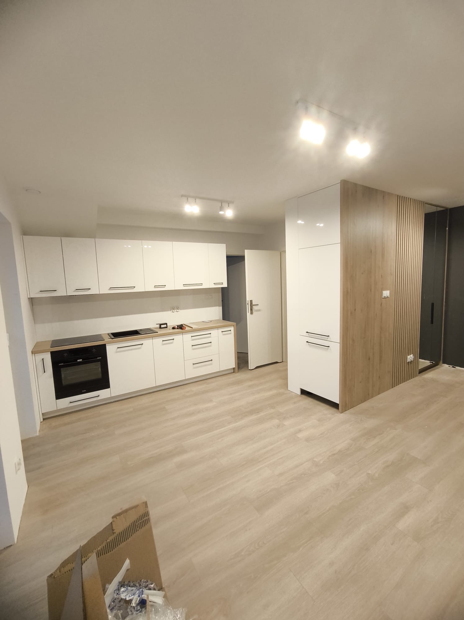 Nowoczesna, jasna kuchnia z białymi szafkami i drewnianym blatem, widok na całe pomieszczenie z częściowo rozpakowanymi materiałami na podłodze.