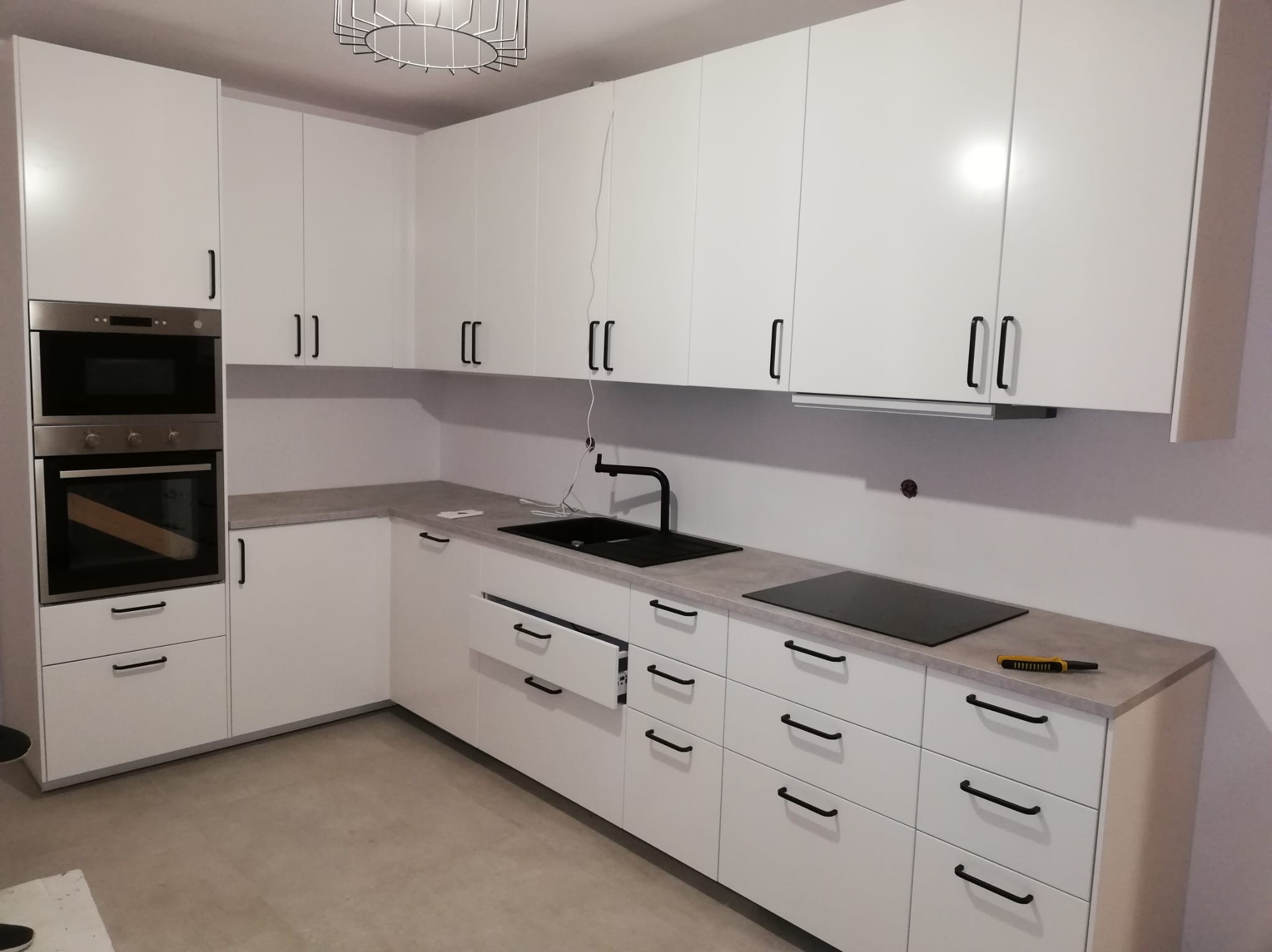 Nowoczesna, biała kuchnia z czarnymi uchwytami, szarym blatem, piekarnikiem w zabudowie, czarną płytą grzewczą i otwartą szufladą, widoczny nożyk na blacie.