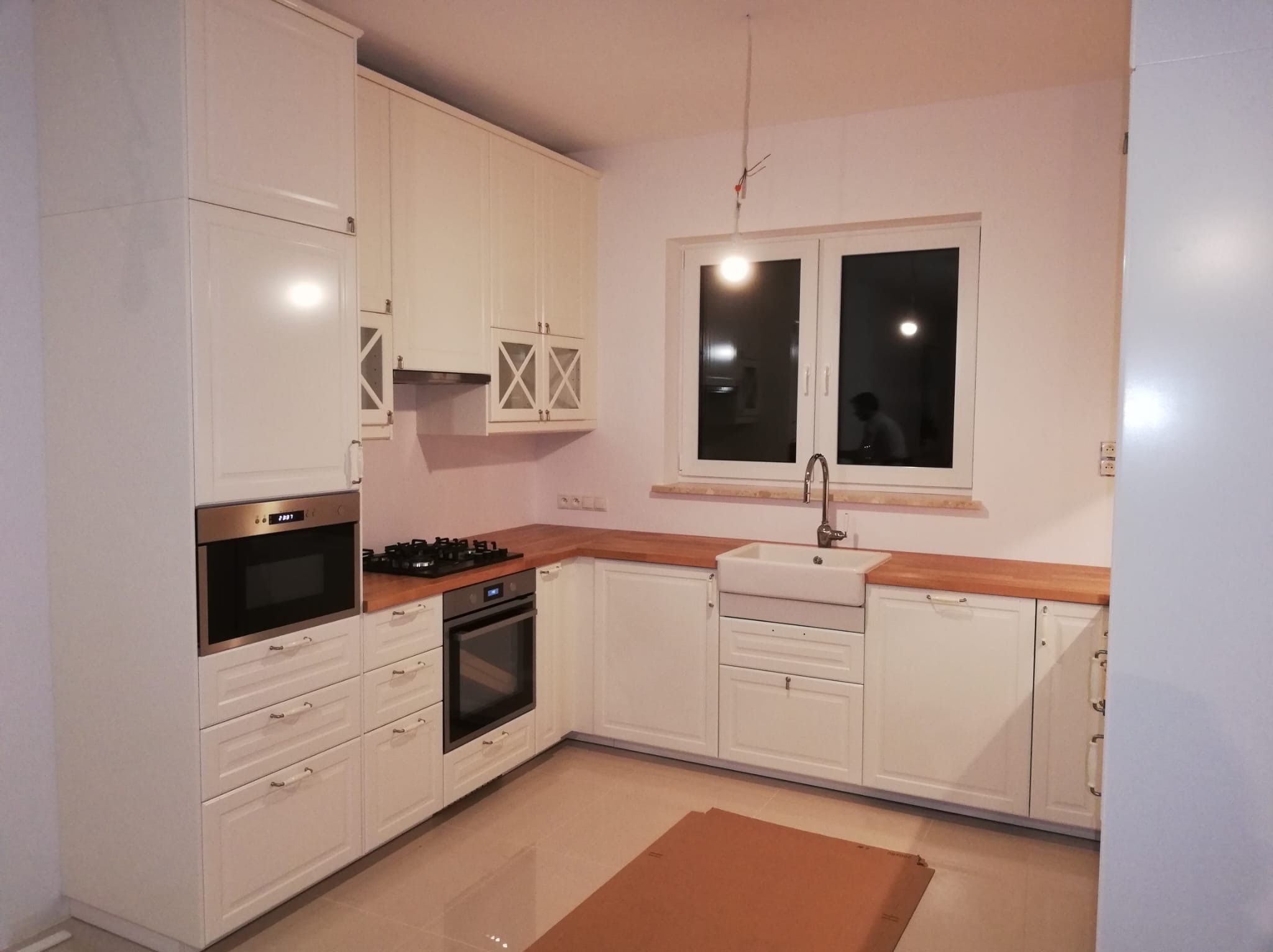 Jasna kuchnia z drewnianym blatem, białą ceramiczną umywalką i wiszącą lampą bez klosza, widok na okno z odbijającą się sylwetką.