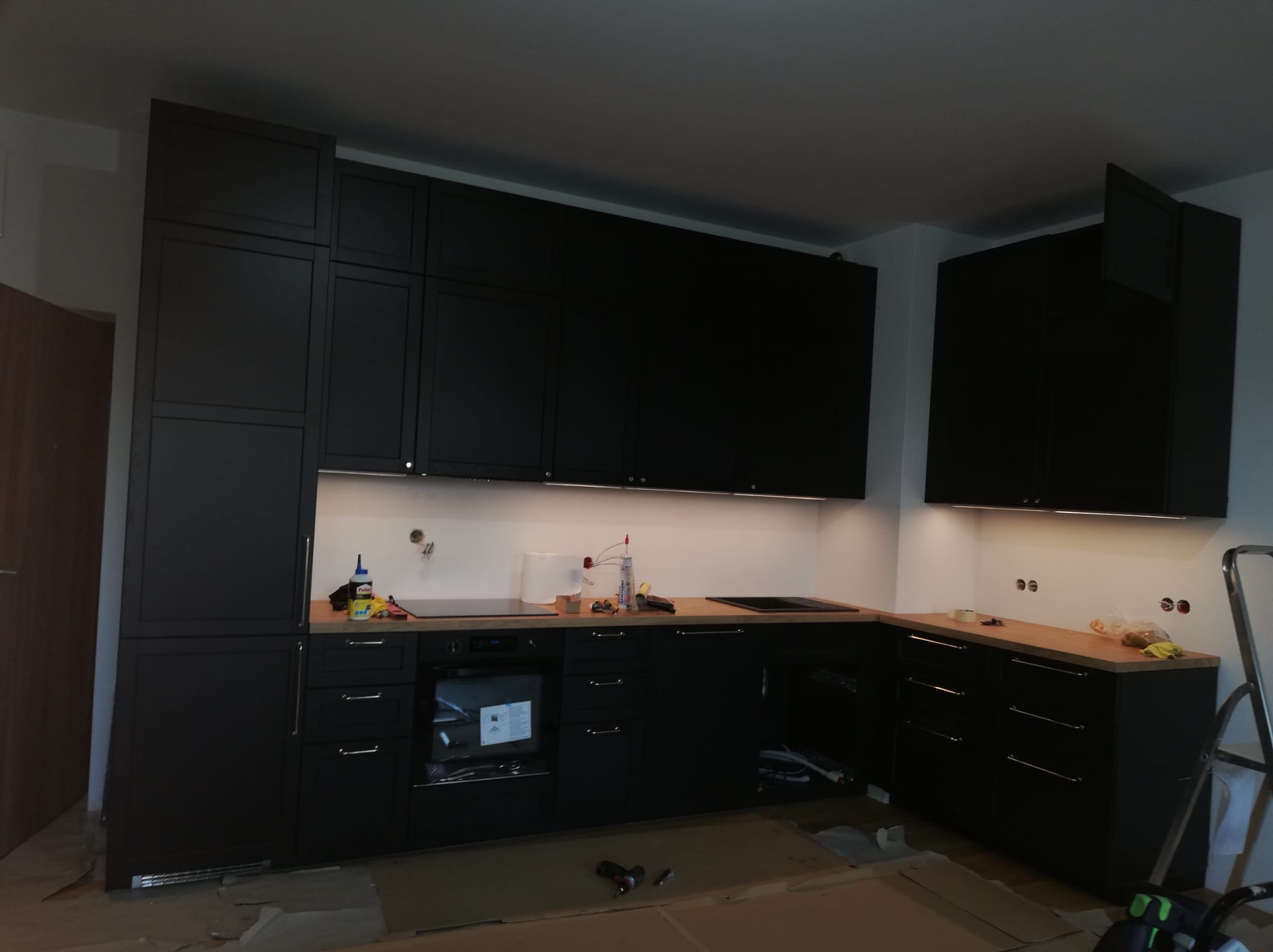 Częściowy widok montażu czarnej zabudowy kuchennej z drewnianym blatem, widoczne narzędzia i kartony na podłodze.