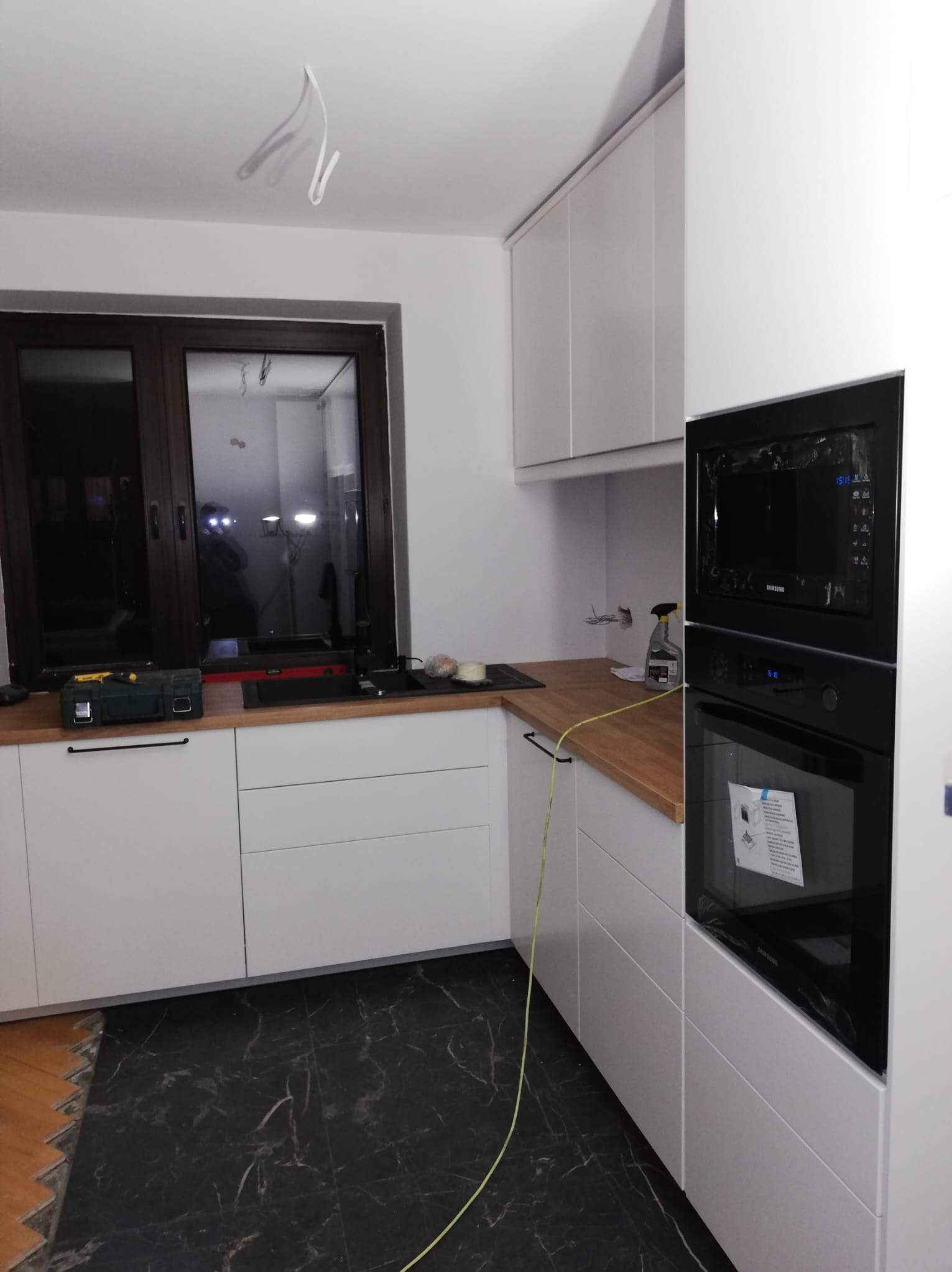 Montaż białej zabudowy kuchennej z drewnianym blatem i czarnym piekarnikiem w trakcie prac wykończeniowych; widoczne narzędzia, niedokończona podłoga i wystające kable.