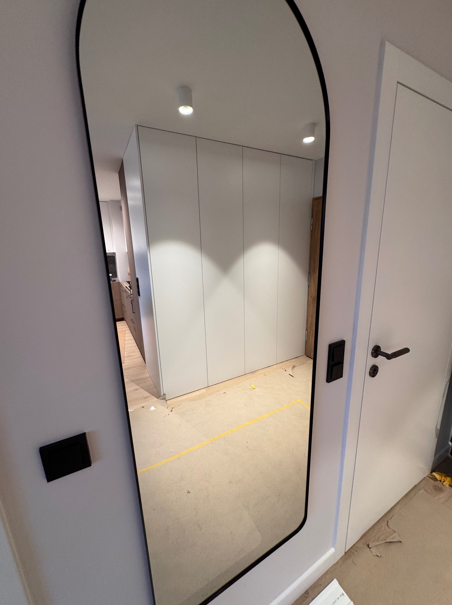 Wnętrze mieszkania z widoczną szafą wnękową w lustrze o nieregularnym kształcie. Obok białe drzwi z czarną klamką. Na ścianie widoczny włącznik światła.