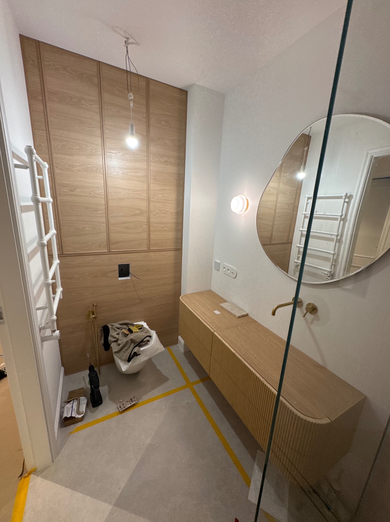 Nowoczesna łazienka w trakcie wykończenia: drewniana zabudowa ściany, szafka pod umywalkę z frezowanym frontem, okrągłe lustro. Widoczne narzędzia i taśmy malarskie.