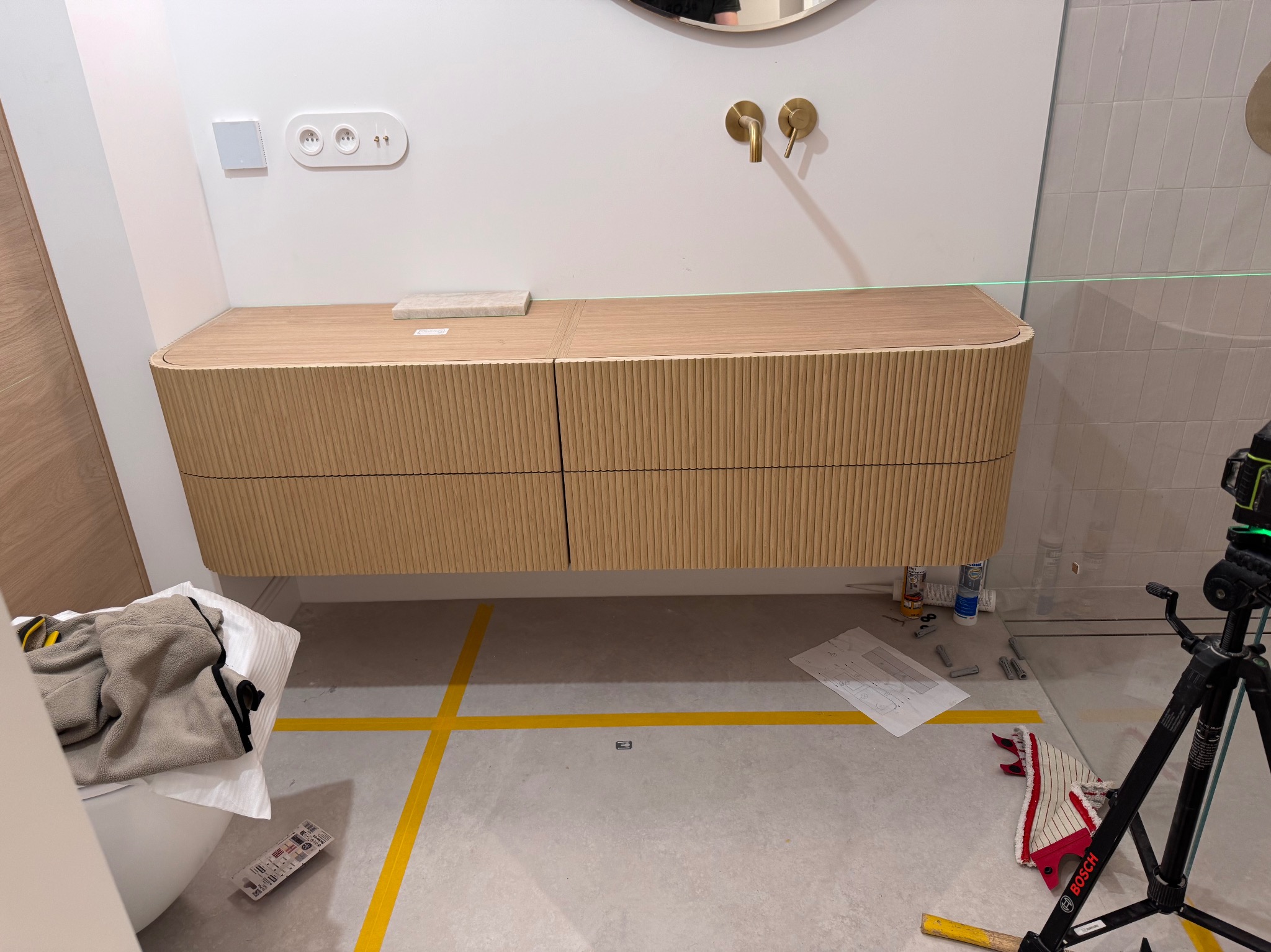 Montaż szafki łazienkowej z drewnianymi frontami, poziomowana laserowo. Widoczne akcesoria montażowe i plan. Umywalka z baterią w kolorze złotym.