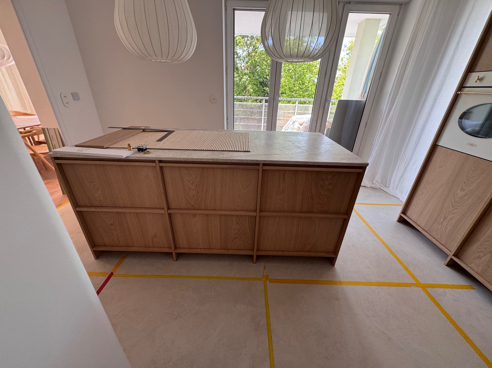 Drewniana wyspa kuchenna z marmurowym blatem w jasnym wnętrzu z widokiem na zewnątrz. Widoczne narzędzia i elementy montażowe na blacie. Żółte taśmy wyznaczają obszar roboczy.