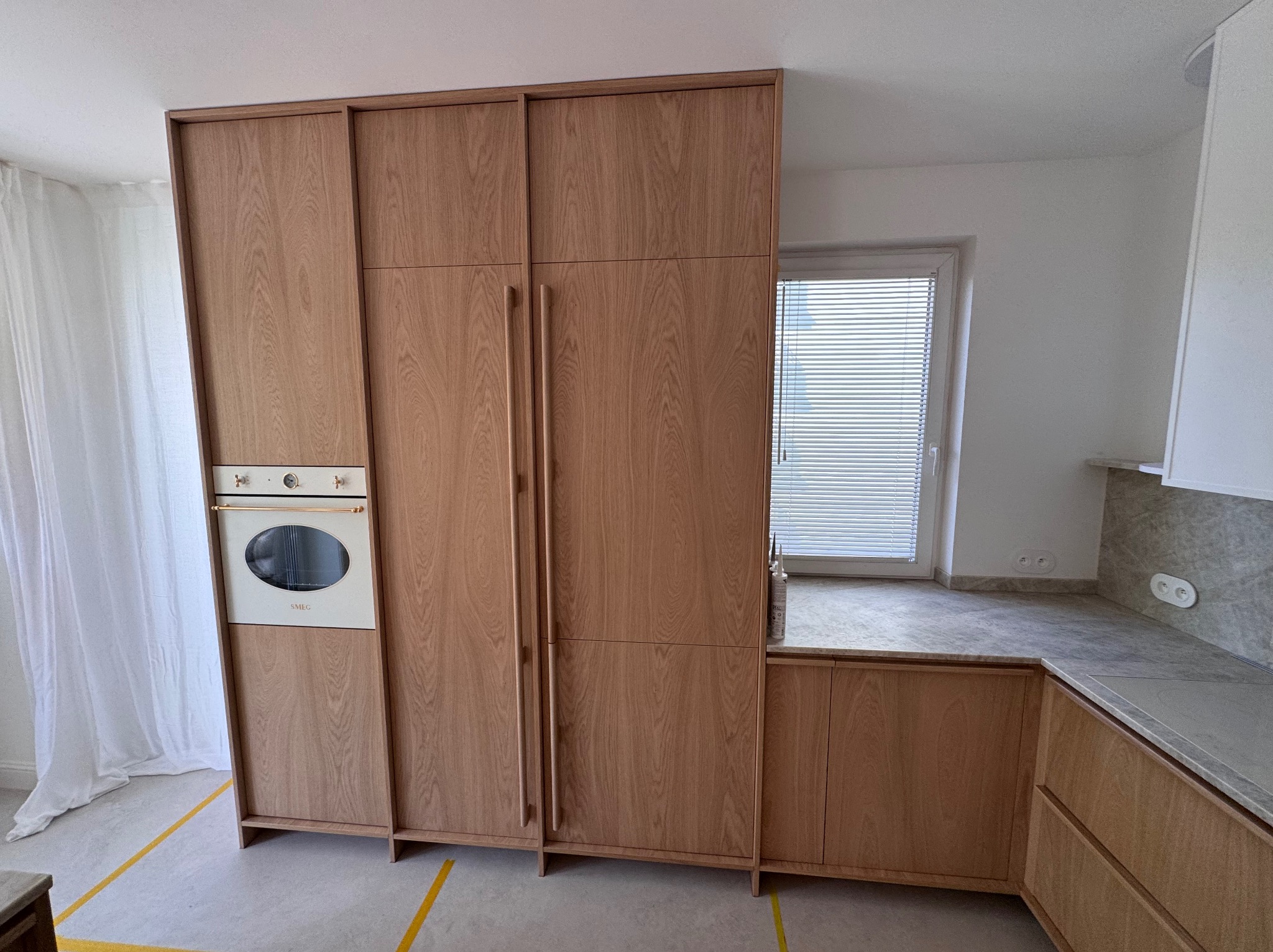 Drewniana zabudowa kuchenna z piekarnikiem SMEG, szare blaty i okno z żaluzjami w tle. Uwagę zwraca naturalne wykończenie drewna i minimalistyczny design.