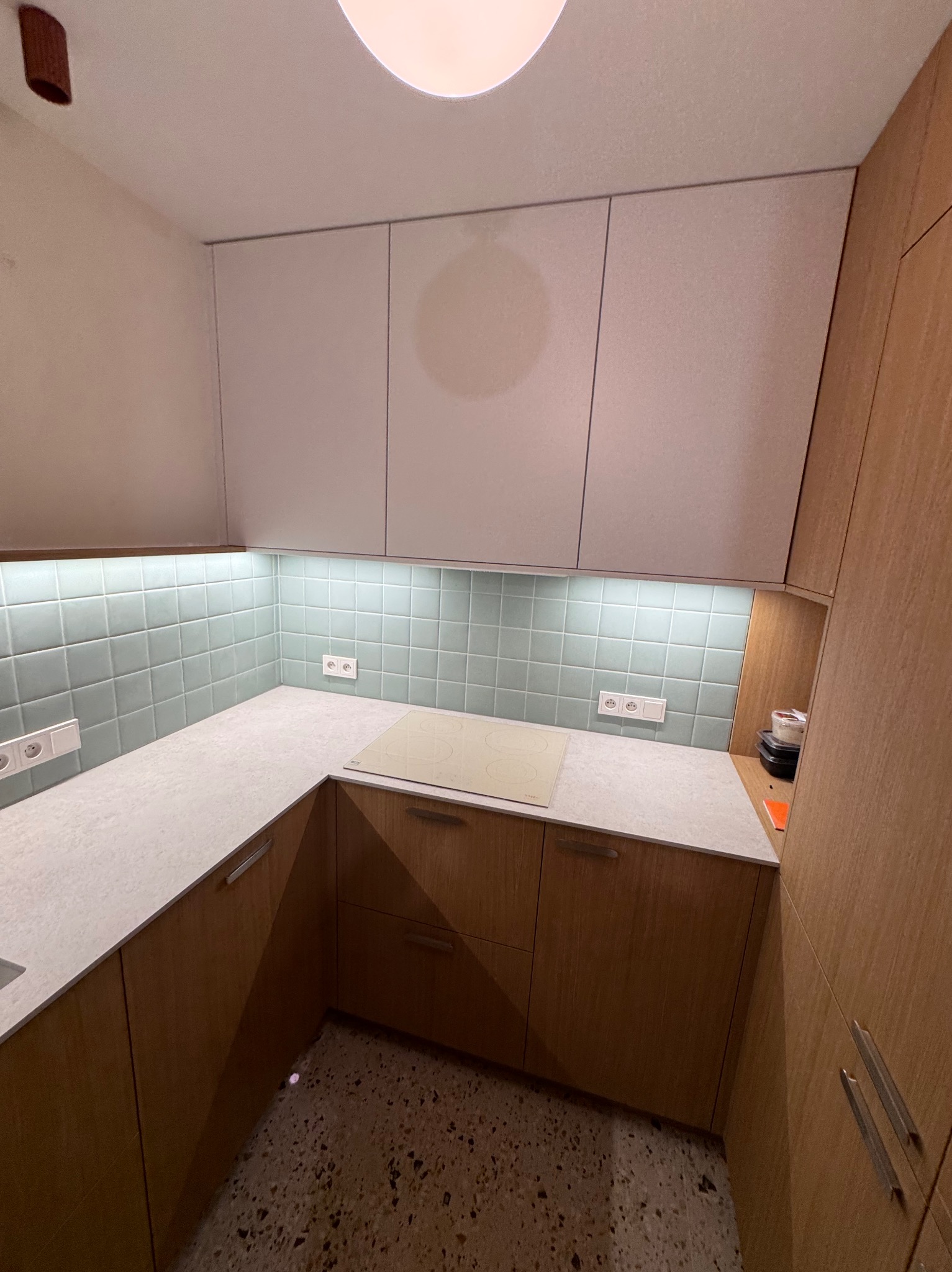 Jasna kuchnia z drewnianymi frontami, białym blatem i pastelowymi płytkami. Widoczne gniazdka elektryczne i płyta grzewcza. Oświetlenie podszafkowe.