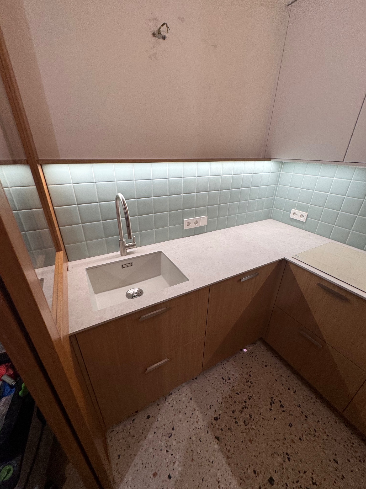 Jasna kuchnia z drewnianymi szafkami, białym blatem i zlewozmywakiem. Nad blatem płytki w odcieniach błękitu i białe gniazdka. Widoczna płyta indukcyjna i oświetlenie podszafkowe.