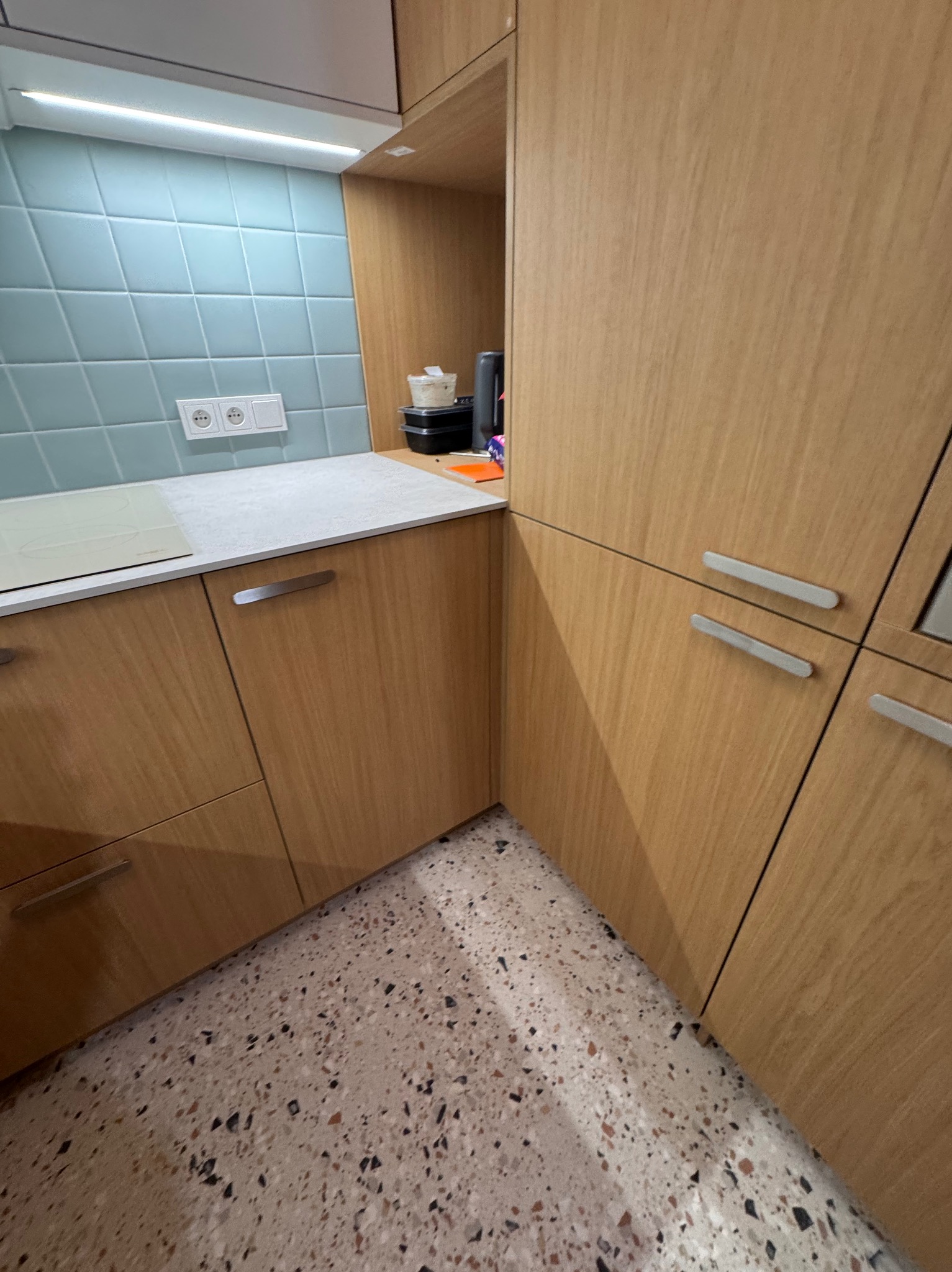 Jasne, drewniane szafki kuchenne z poziomymi, metalowymi uchwytami, błękitne płytki na ścianie i podłoga lastryko. Widok na fragment zabudowy kuchennej.