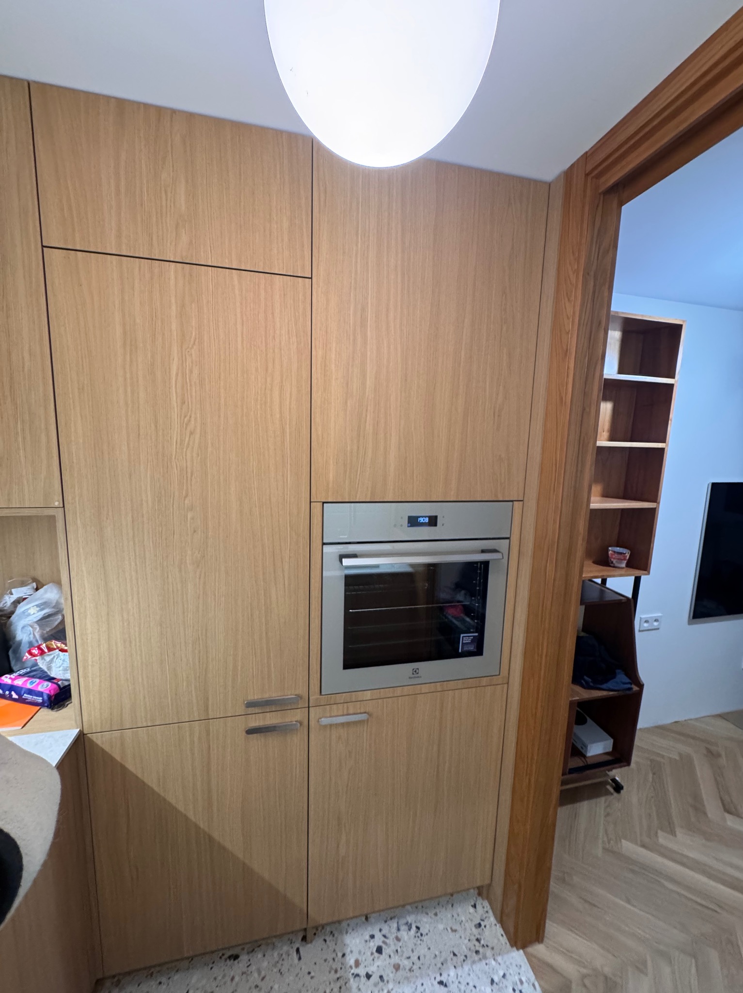 Jasna, drewniana zabudowa kuchenna z piekarnikiem wkomponowanym w szafkę. Widoczny fragment blatu i podłogi. W tle regał i telewizor.