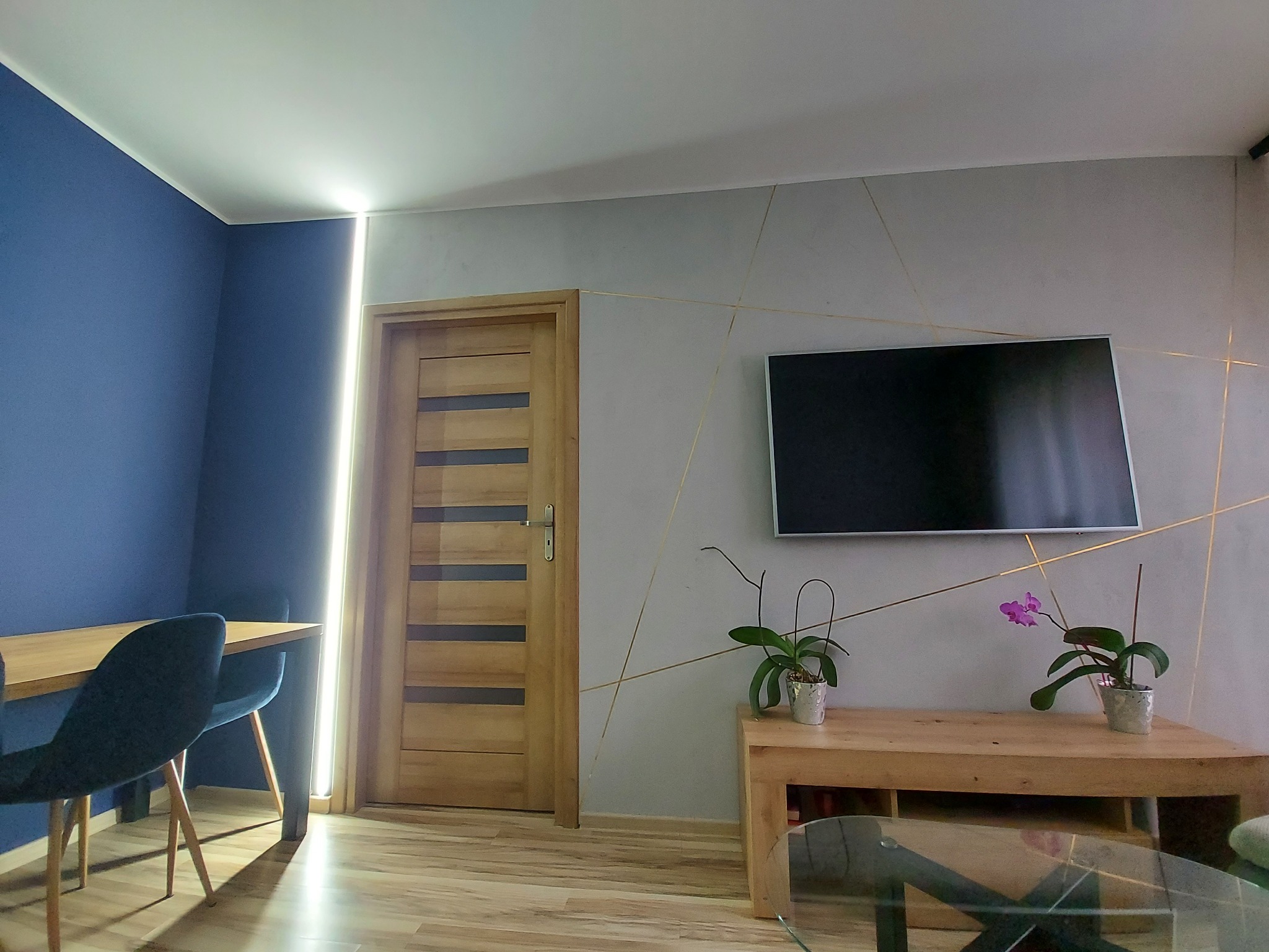 Nowoczesne wnętrze: granatowa ściana, szara ściana z geometrycznymi złotymi liniami, drewniane drzwi z przeszkleniami, telewizor, drewniana konsola z dwoma storczykami, stół z krzesłami.