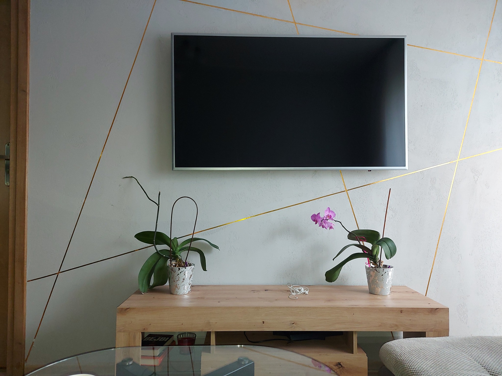 Ściana w salonie z dekoracyjnym szpachlowaniem w geometryczne złote linie, podwieszony telewizor, drewniana szafka z dwoma doniczkami z orchideami.
