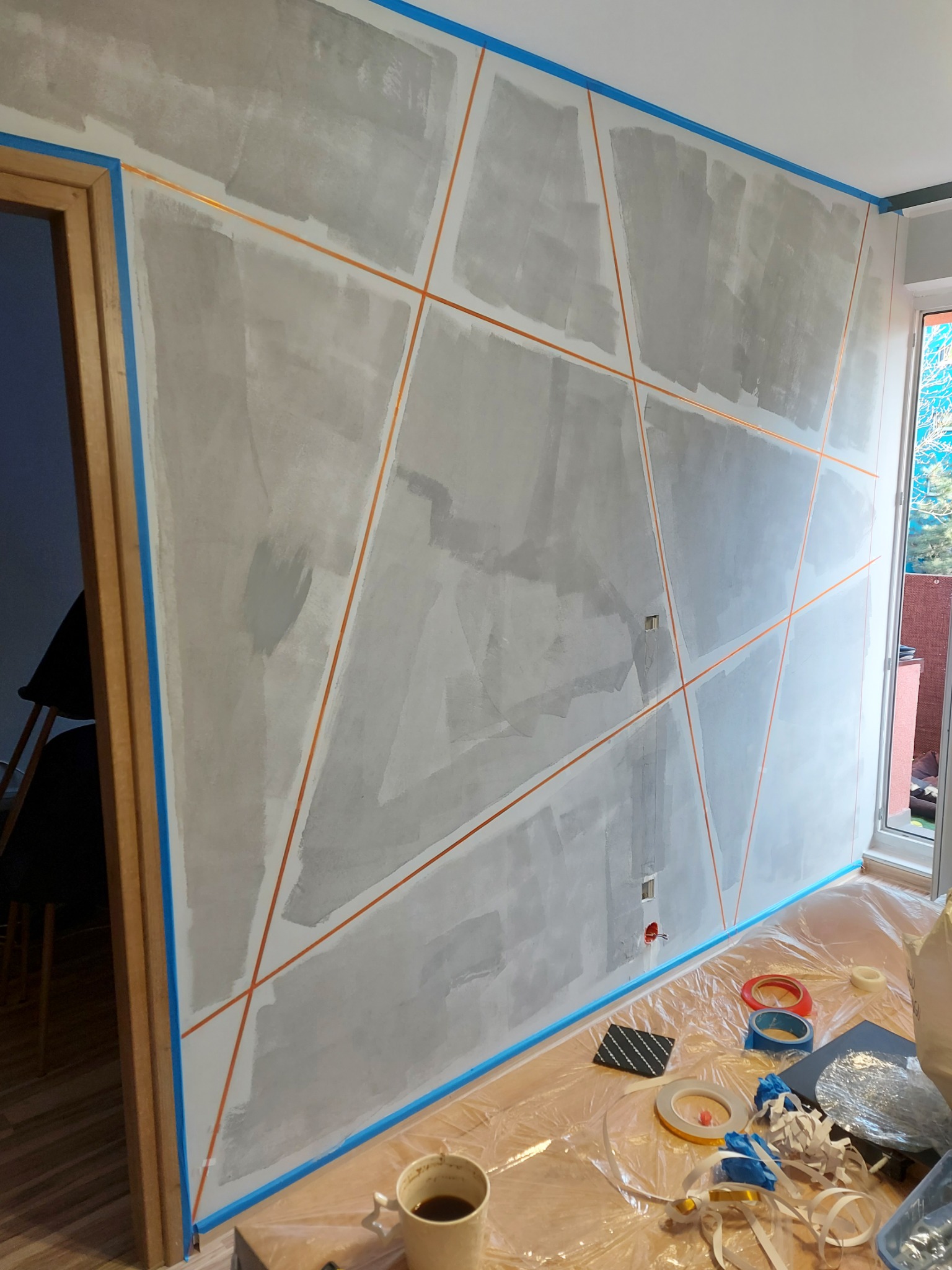 Przygotowanie ściany do malowania w Zabrzu: ściana z nałożoną warstwą szpachli, wyklejona taśmą malarską w geometryczne wzory, widoczne narzędzia i zabezpieczenia podłogi.