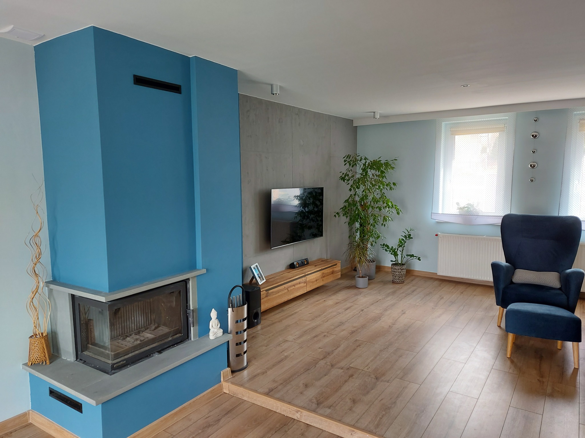 Salon po remoncie: błękitny kominek z szarym obramowaniem, drewniana podłoga, szara ściana z telewizorem, fotel w odcieniach granatu, roślina doniczkowa i dekoracje.