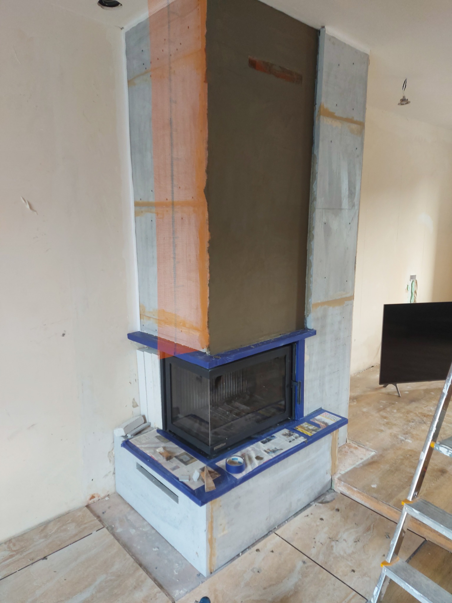 Wnętrze w trakcie budowy kominka: ściana z siatką i nałożoną zaprawą, obudowa kominka zabezpieczona niebieską taśmą malarską, widoczna drabina i niedokończona podłoga.