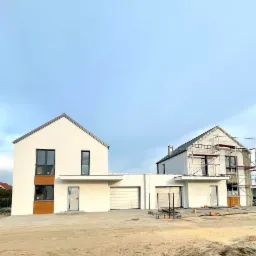 Dwa nowoczesne, białe domy jednorodzinne w trakcie budowy, jeden z elewacją częściowo wykończoną i stolarką okienną, drugi z rusztowaniami i nieukończoną elewacją, na piaszczystym placu budowy.