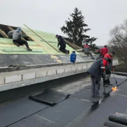 Prace dekarskie na dachu: ekipa montuje papę termozgrzewalną przy użyciu palników gazowych, widoczna butla z gazem i przygotowane rolki papy, w tle konstrukcja drewniana dachu i drzewo