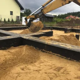 Żółta koparka wypełnia piaskiem czarne, zabezpieczone fundamenty domu na działce z widocznymi budynkami w tle.