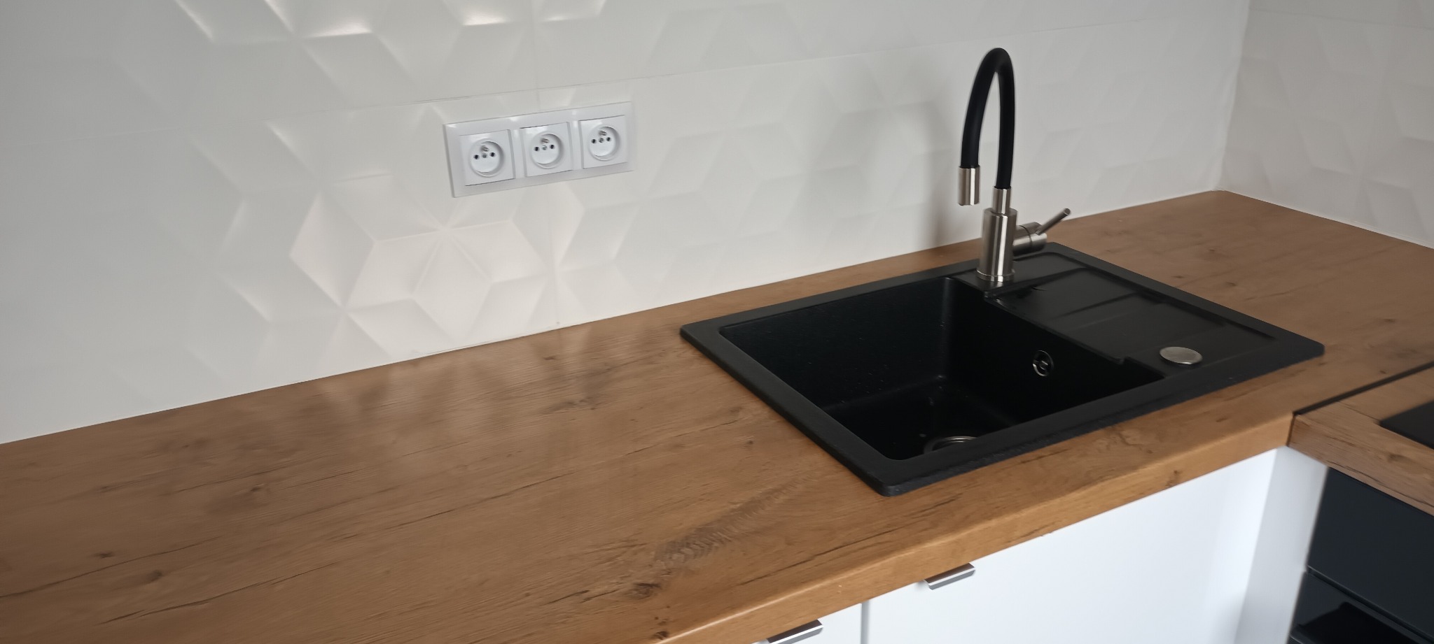 Nowoczesna kuchnia: czarny zlew granitowy z chromowaną baterią na blacie w kolorze drewna, białe, trójwymiarowe płytki na ścianie z trzema gniazdkami.