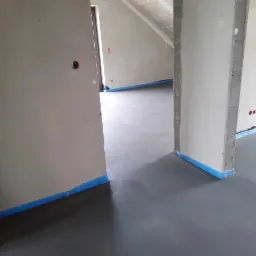 Świeżo wylana, szara posadzka w surowym wnętrzu z widocznymi ścianami przygotowanymi do wykończenia, zabezpieczonymi niebieską taśmą malarską.