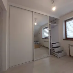 Biała szafa wnękowa z przesuwnymi drzwiami z lustrem, wbudowana w pokoju na poddaszu, z otwartymi półkami i szufladami po prawej stronie.