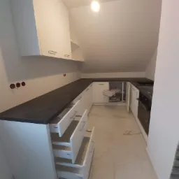 Kuchnia na poddaszu z białymi szafkami i szufladami Blum, ciemnym blatem i karuzelą w szafce narożnej, widoczne niedokończone gniazdka elektryczne.