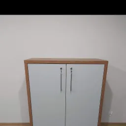 Niska szafka z dwoma białymi drzwiczkami, obramowana drewnem, z długimi, srebrnymi uchwytami, stojąca na podłodze przy białej ścianie z widocznym gniazdkiem elektrycznym.