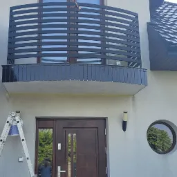 Elewacja domu z czarnym, nowoczesnym balkonem z poziomymi szczeblami, widoczna drabina malarska i ślady świeżego malowania na ścianie pod balkonem.