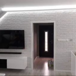 Salon z białą cegłą dekoracyjną na ścianie, oświetleniem LED podsufitowym, wiszącym kryształowym żyrandolem, szafką RTV i telewizorem, widok na otwarte drzwi do innego pomieszczenia.