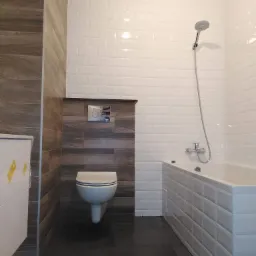 Nowoczesna łazienka z białymi, prostokątnymi płytkami na ścianach i obudowie wanny, kontrastującymi z ciemną podłogą i fragmentem ściany wyłożonym drewnopodobnymi panelami, wisząca toaleta...