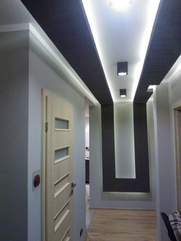 Nowoczesny korytarz z podwieszanym sufitem z oświetleniem LED, drewnianą podłogą i jasnymi ścianami, widoczna drewniana drzwi z przeszkleniami.