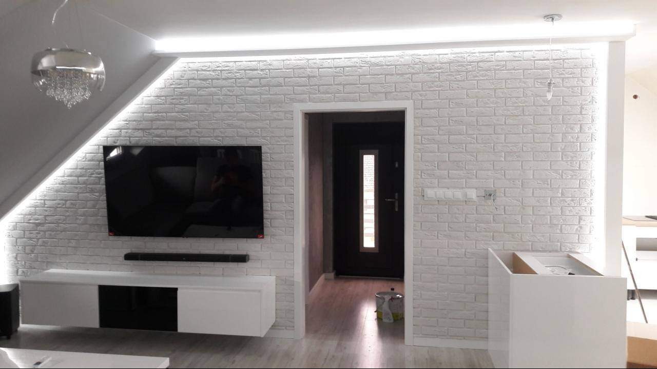 Salon z białą cegłą dekoracyjną na ścianie, oświetleniem LED podsufitowym, wiszącym kryształowym żyrandolem, szafką RTV i telewizorem, widok na otwarte drzwi do innego pomieszczenia.