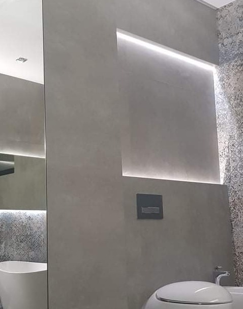 Nowoczesna łazienka z szarą, betonową ścianą z podświetlaną wnęką, białą ceramiką i dekoracyjną mozaiką po prawej stronie.