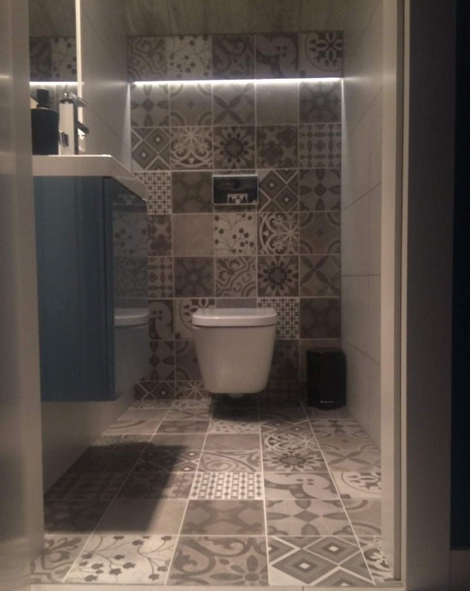 Toaleta w małej łazience z patchworkowymi płytkami podłogowymi i ściennymi w odcieniach szarości, podświetlona listwą LED nad muszlą, obok wisząca szafka umywalkowa w kolorze niebieskim.