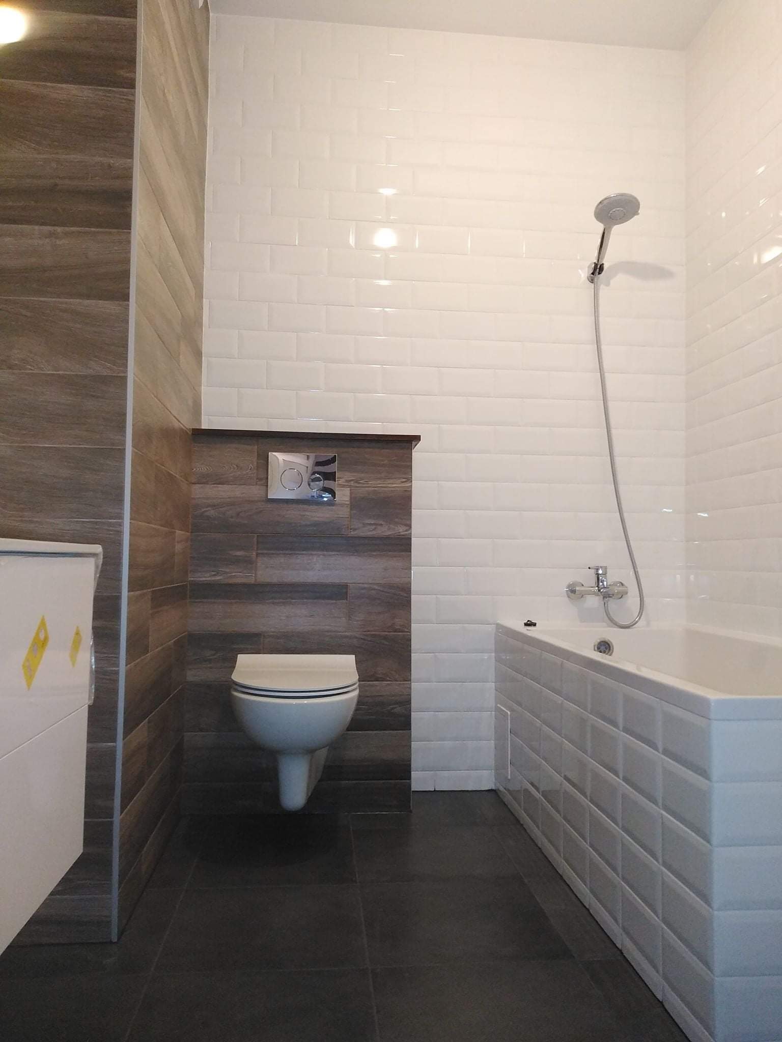 Nowoczesna łazienka z białymi, prostokątnymi płytkami na ścianach i obudowie wanny, kontrastującymi z ciemną podłogą i fragmentem ściany wyłożonym drewnopodobnymi panelami, wisząca toaleta...