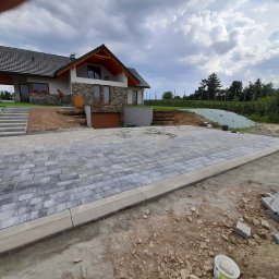 MAXBRUK-BUD - Ułożona szara kostka brukowa na podjeździe przed nowoczesnym domem z brązowymi akcentami, widoczne narzędzia i resztki kostki brukowej na ziemi, pracownik w tle.