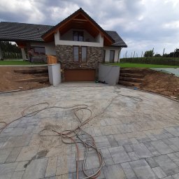 MAXBRUK-BUD - Ułożona szara kostka brukowa na podjeździe przed nowoczesnym domem z garażem, widoczne kable i prace ziemne w tle.