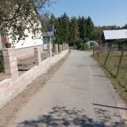 Nowo wybudowany, beżowy, modułowy płot z kamiennych bloczków, oddzielający posesję od drogi dojazdowej, w tle dom i drzewa.