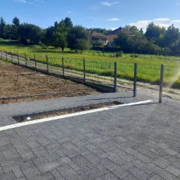 Nowo ułożona, szara kostka brukowa z białym pasem oddzielającym od rabaty. Widoczny fragment ogrodzenia i teren przygotowany pod nasadzenia. W tle domy i zieleń.