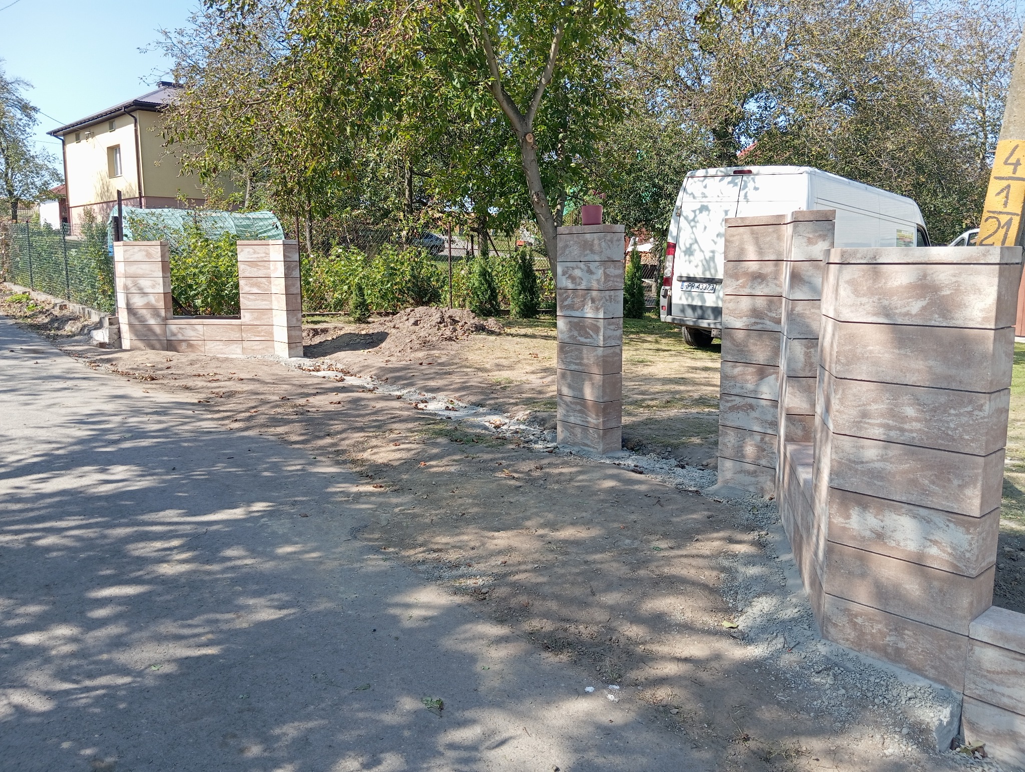 Wznoszenie ogrodzenia z kostki imitującej kamień w kolorach beżu i brązu, widoczny biały van w tle, teren w trakcie prac ziemnych.