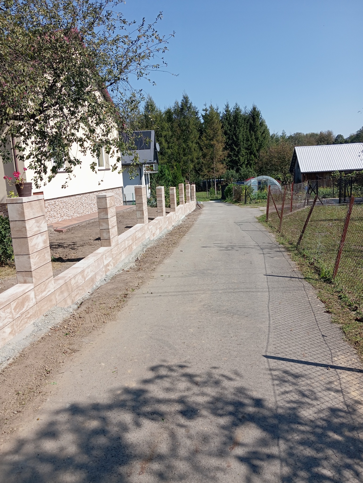 Nowo wybudowany, beżowy, modułowy płot z kamiennych bloczków, oddzielający posesję od drogi dojazdowej, w tle dom i drzewa.