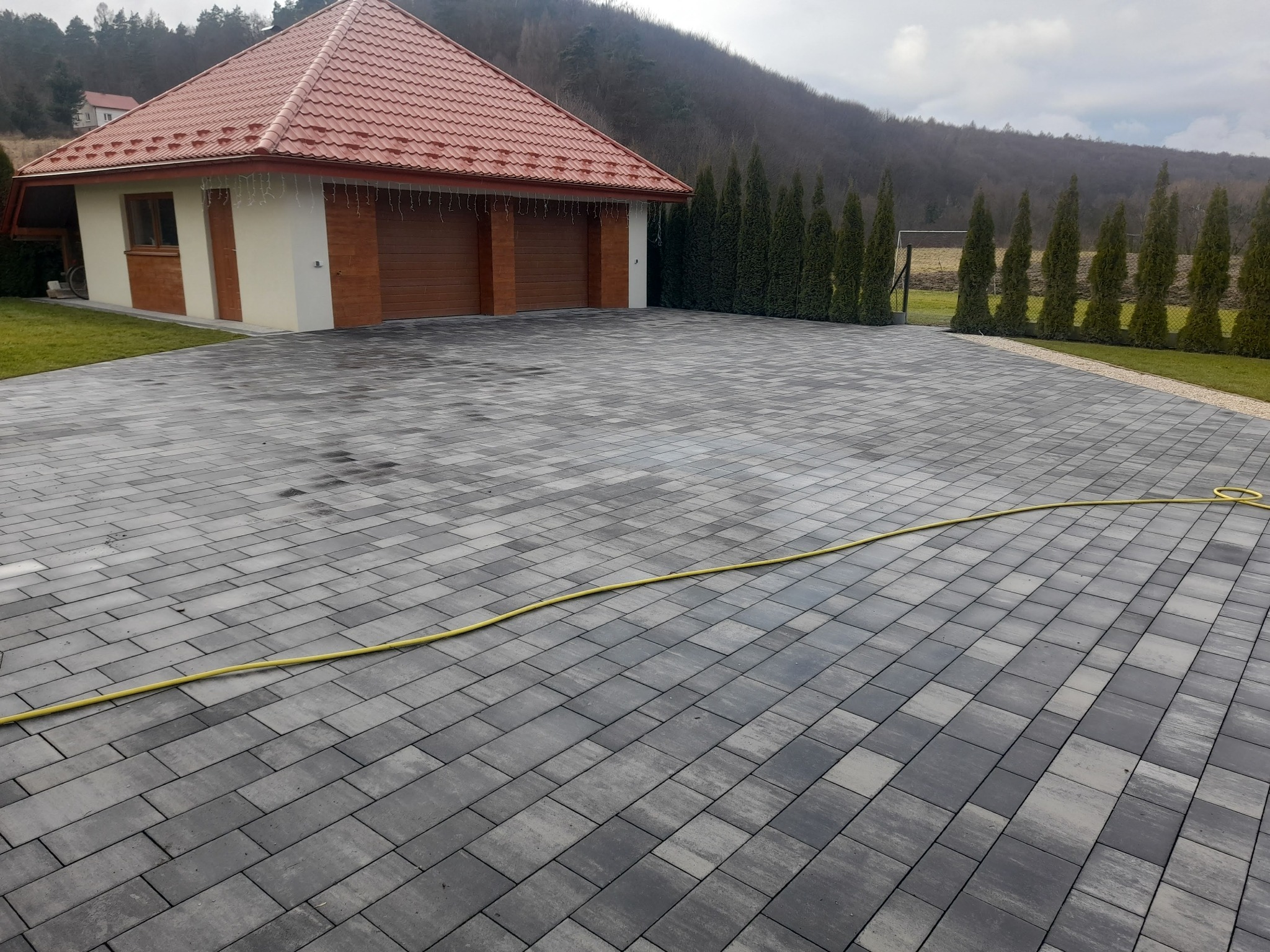 Nowa, szara nawierzchnia z kostki brukowej przed domem z dwustanowiskowym garażem i rzędem tuj, z widocznym żółtym wężem ogrodowym.