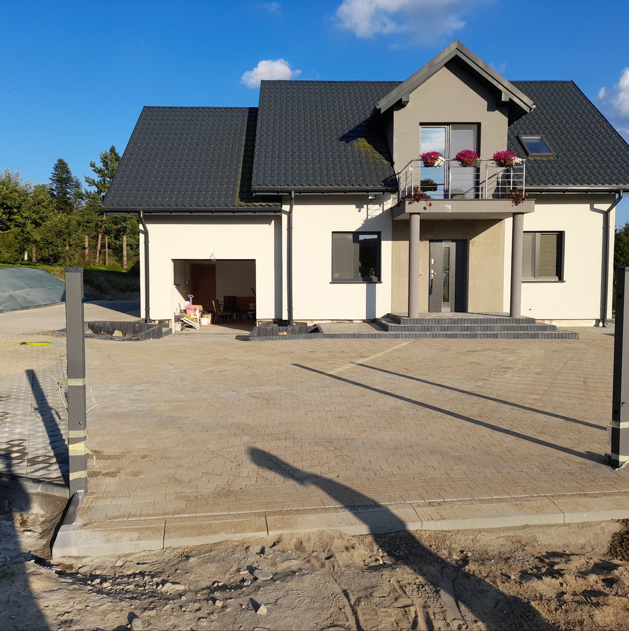 Nowoczesny dom jednorodzinny z szarą kostką brukową na podjeździe, widoczne przygotowania do montażu ogrodzenia.