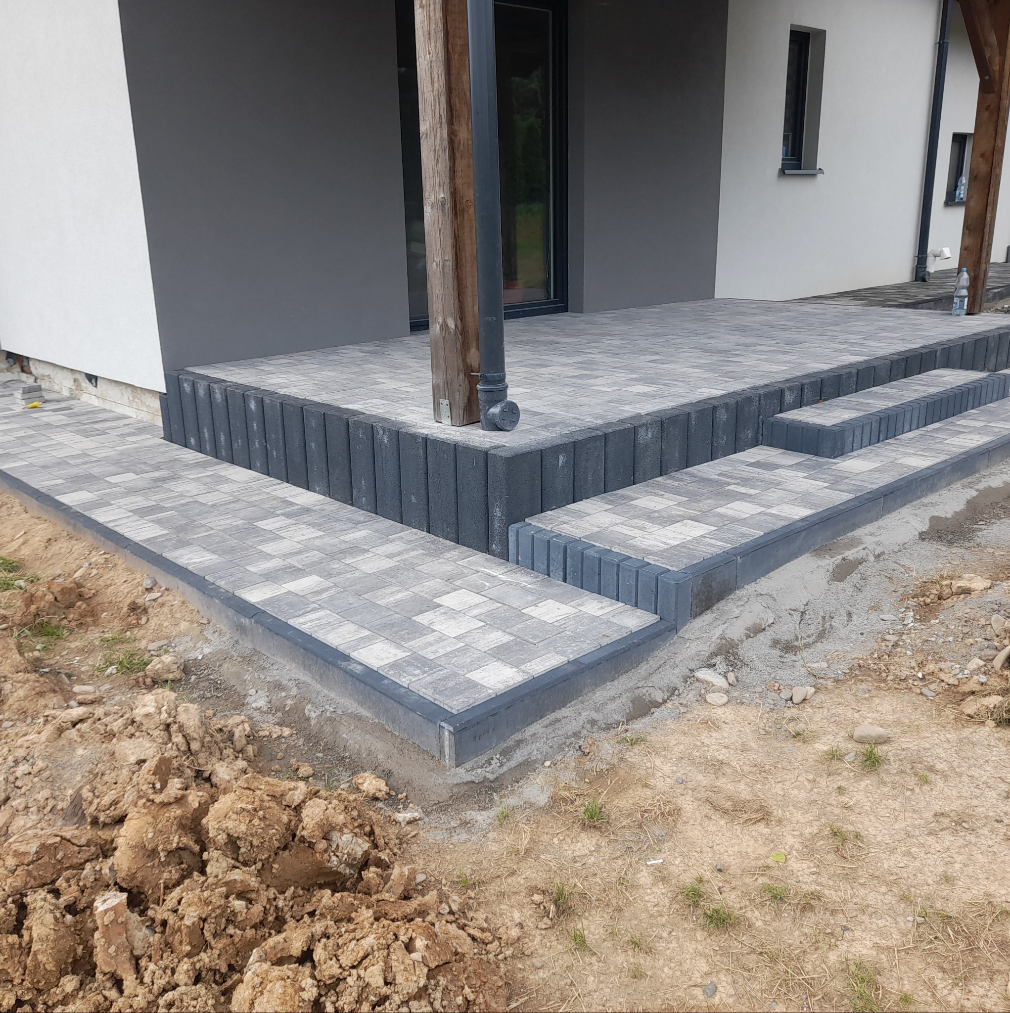Schody wejściowe do domu wyłożone szarą kostką brukową, obrzeża z pionowo ułożonych elementów, widoczne ślady prac ziemnych.
