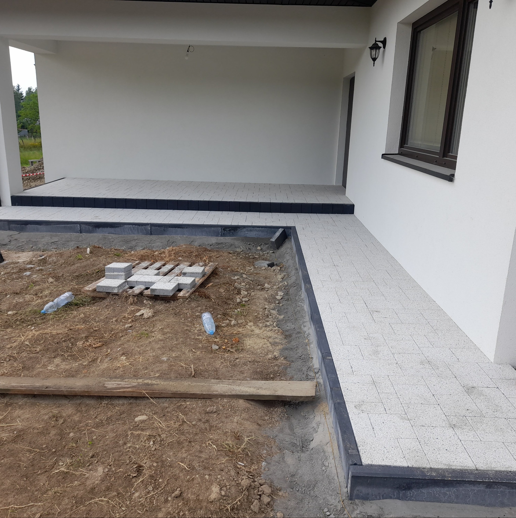 Fragment posesji z częściowo ułożoną jasną kostką brukową, widoczne przygotowane podłoże, paleta z kostką i butelki po wodzie. Czarny obrzeże. Dom z białymi ścianami w tle.