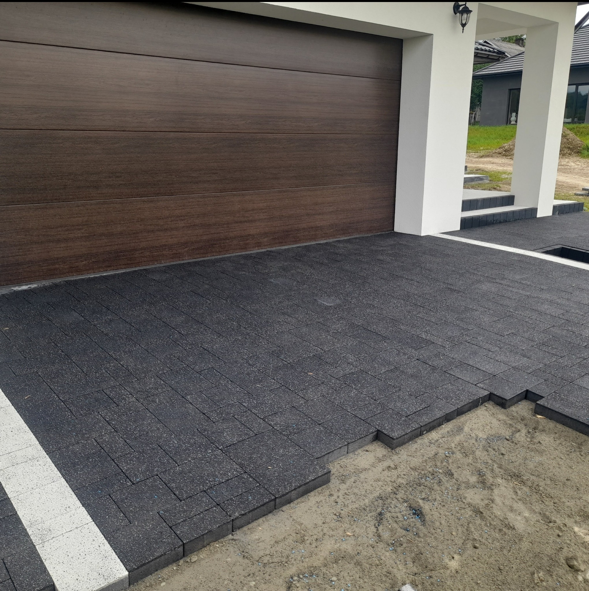 Niedokończona nawierzchnia z grafitowej kostki brukowej, widoczne podsypka piaskowa i fragment garażu z brązową bramą.